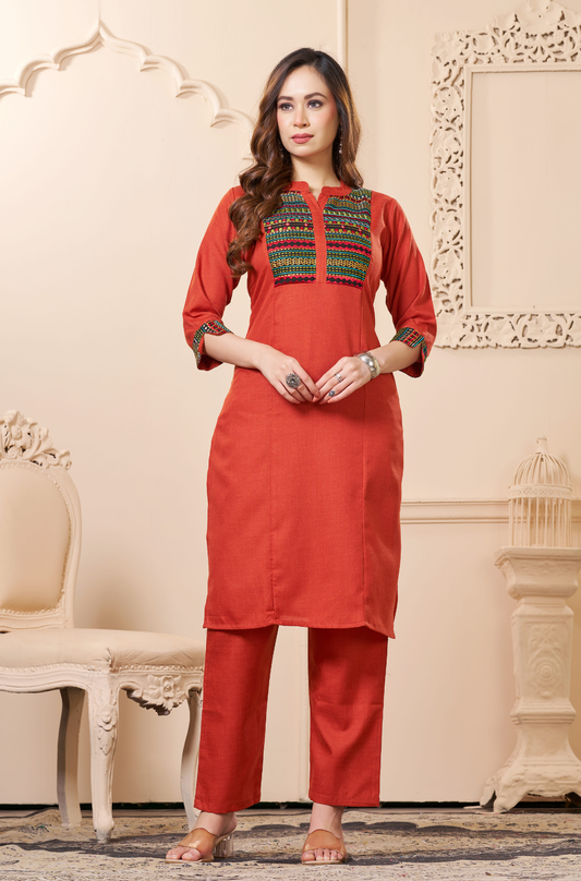 A woman standing in a rust-orange straight kurta featuring a multi-colored geometric embroidered yoke  and matching straight-leg trousers.