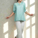 Front view of women's sea green rayon Top featuring a mandarin collar, button-down front, front pleats, half sleeves, and a relaxed fit paired with white trousers.
