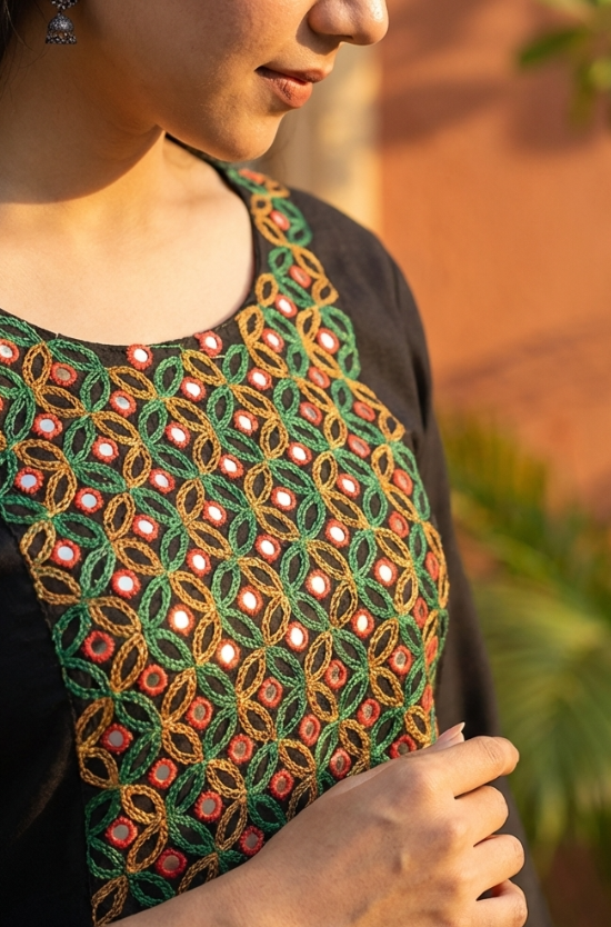 A close-up view of the neckline of a black kurta, highlighting detailed green, red, and gold mirror-work embroidery.