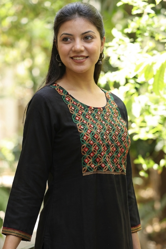 A portrait of a woman wearing a black ethnic kurta with mirror-work, set against a blurred green background.