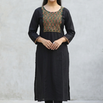A woman leaning against a stone wall, wearing a black straight kurta with mirror-work embroidery.