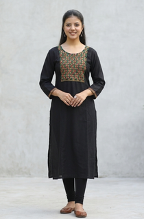 A woman leaning against a stone wall, wearing a black straight kurta with mirror-work embroidery.