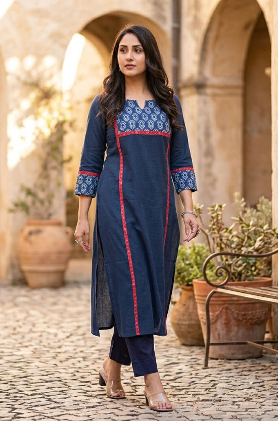 Woman walking outdoors on a cobbled path wearing a stylish blue kurta with red panel borders and dark blue trousers.