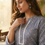 A woman standing sideways, modeling a grey-blue straight-cut kurta with white lace trim and white trousers.
