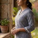 A side-profile portrait of a woman standing near a stone wall, modeling a grey-blue printed ethnic kurta.