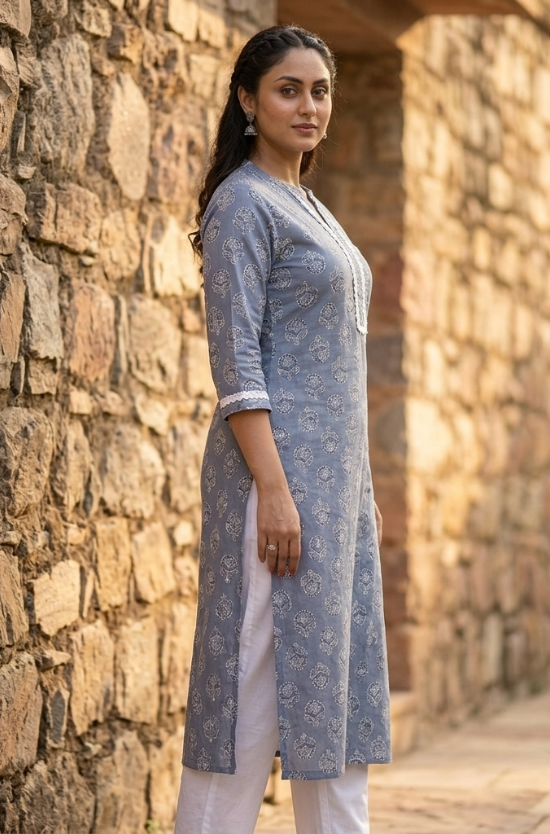 A side-profile portrait of a woman standing near a stone wall, modeling a grey-blue printed ethnic kurta.