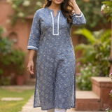 A woman walking gracefully in a garden wearing a grey-blue floral printed kurta and white straight pants.