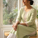 A woman sitting gracefully by a window wearing a light green cotton kurta with pink floral embroidery and white palazzo pants.