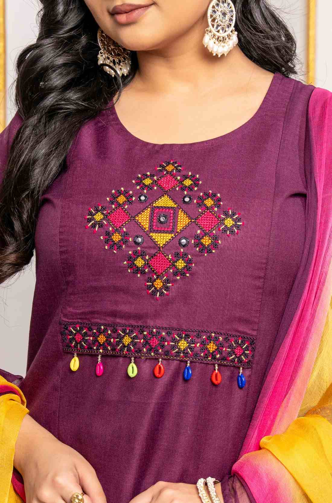Close-up of multicolour geometric cross stitch embroidery, mirror accents and decorative tassels on magenta cotton kurta.