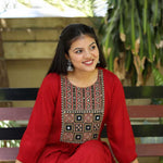 A model sitting on a bench while wearing a maroon rayon round-neck A-line kurta with 3/4 sleeves and a block-printed embroidered yoke