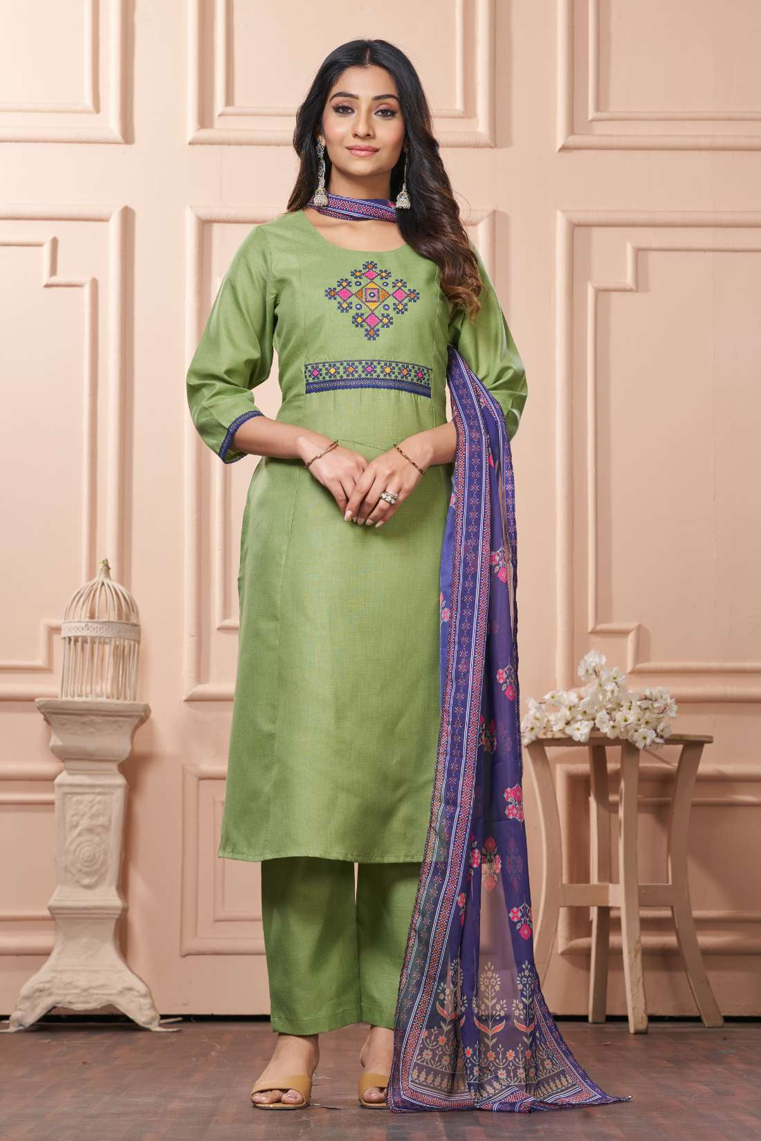 A full length presentation of a pastel green cotton ethnic suit set displaying vibrant floral cross stitch embroidery details along the front panel, styled with straight pants and a printed dupatta