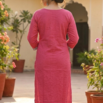 A woman standing with her back to the camera, wearing a pink textured kurta with white pants in a garden.