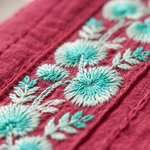 A detailed macro close-up of a pink cotton kurta showing intricate turquoise and white floral thread embroidery work.