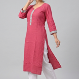 A woman posing sideways wearing a pink embroidered kurta and white trousers against a neutral background.