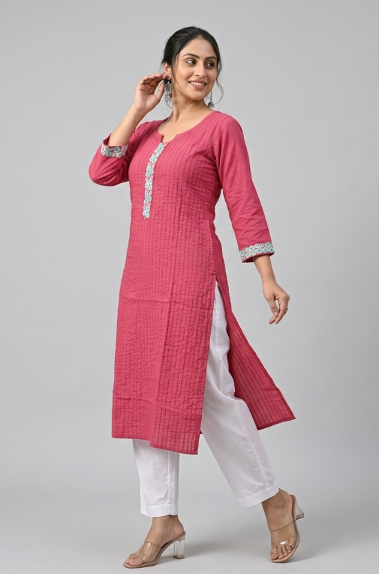 A woman posing sideways wearing a pink embroidered kurta and white trousers against a neutral background.