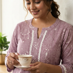 A lifestyle shot of a smiling woman holding a tea cup, wearing a purple ethnic kurta with lace trim.
