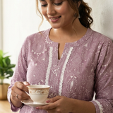 A lifestyle shot of a smiling woman holding a tea cup, wearing a purple ethnic kurta with lace trim.