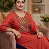A woman sitting on a sofa, modeling a rust-orange ethnic kurta with detailed embroidery and blue bottoms.