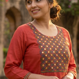 A woman smiling, wearing a rust-orange straight kurta with intricate ethnic embroidery on the chest.
