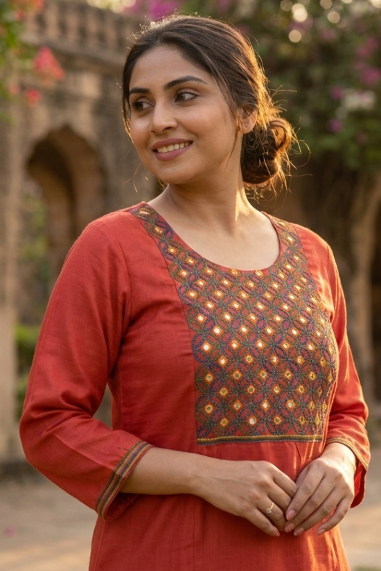 A woman smiling, wearing a rust-orange straight kurta with intricate ethnic embroidery on the chest.