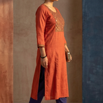 A woman standing sideways, modeling a rust-orange straight-cut ethnic kurta with blue trousers.
