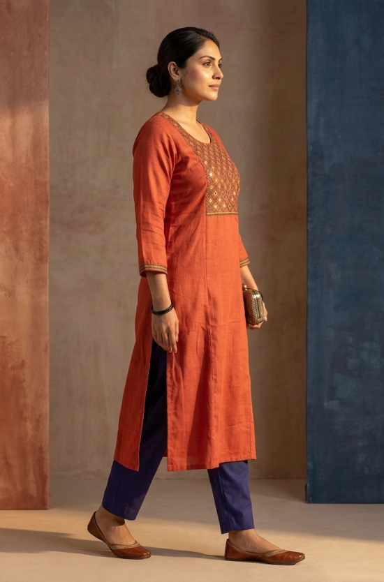 A woman standing sideways, modeling a rust-orange straight-cut ethnic kurta with blue trousers.