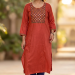 A woman walking gracefully in a garden wearing a rust-orange ethnic kurta and blue trousers.