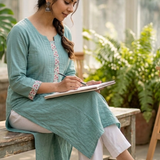 A woman sitting on a wooden bench sketching in a notebook, wearing a sea-green embroidered kurta.