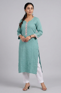 A full-length studio shot of a woman modeling a sea-green straight kurta with white pants. A woman posing with her hands near her face, wearing a sea-green embroidered kurta in a garden sea-green-kurta-walking-shot. A woman walking in a garden wearing a sea-green kurta, show casing the flow and drape of the fabric. sea-green-kurta-tea-lifestyle. A woman holding a clay cup while leaning against a wooden gate, dressed in a sea-green ethnic kurta.