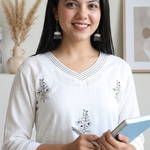 A smiling woman holding a notebook and pen, wearing a white block-printed ethnic kurta.