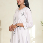 A woman standing in a white straight-cut kurta with detailed black floral patterns and sleeve stitching.