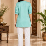 Full-length back view of a woman wearing the light sea green rayon pleated top paired with white pants.