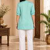 Full-length back view of a woman wearing the light sea green rayon pleated top paired with white pants.