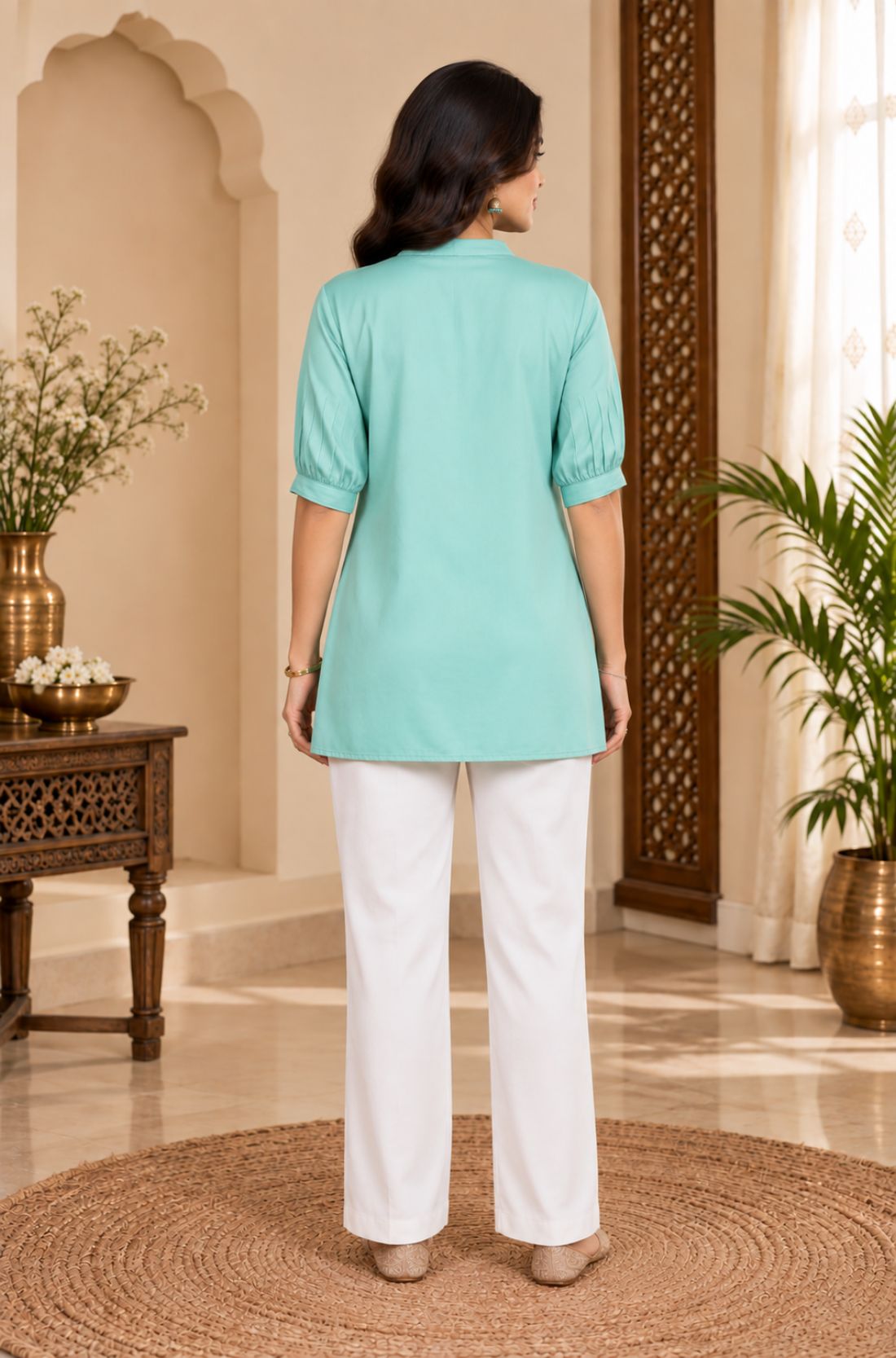 Full-length back view of a woman wearing the light sea green rayon pleated top paired with white pants.