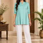 Full-length front view of a model wearing a light sea green rayon pleated top with white trousers.